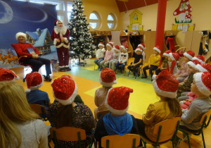 Grupa dzieci siedzi na krzesłach wokół pana Marka Cieślaka, który czyta dzieciom książkę. W tle dekoracja świąteczna.